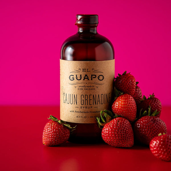 Bottle of large Guapo Cajun Grenadine syrup with strawberries on a red surface and pink background