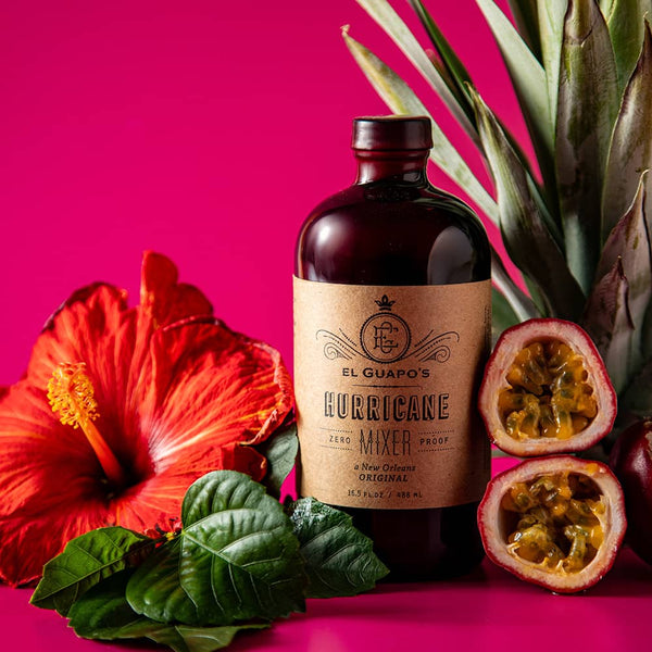 Bottle of full size El Guapo Hurricane Mixer with tropical flowers and fruit on a pink background