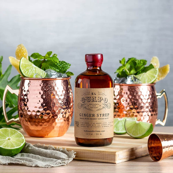 Bottle of El Guapo Ginger Syrup with copper mugs and garnishes on a wooden board.