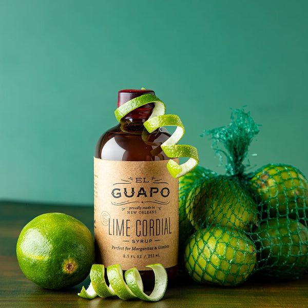 Bottle of small 8.5 oz El Guapo Lime Cordial Syrup with limes on a green background