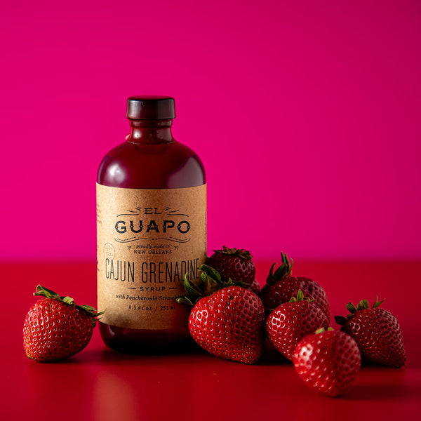 8.5 oz Bottle of Guapo Cajun Grenadine syrup with strawberries on a red surface and pink background