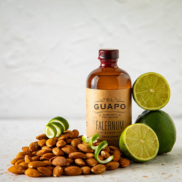 Bottle of Small El Guapo Falernum syrup with almonds and limes on a light background