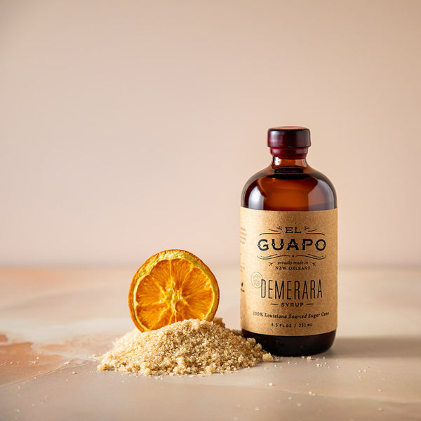 Small glass bottle of El Guapo Demerara syrup with a dried orange slice and sugar on a beige background