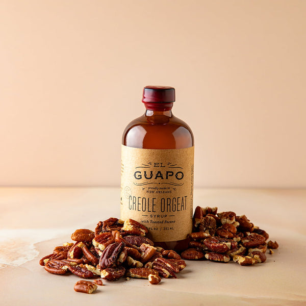 Small bottle of El Guapo Creole Orange syrup surrounded by pecans on a beige background