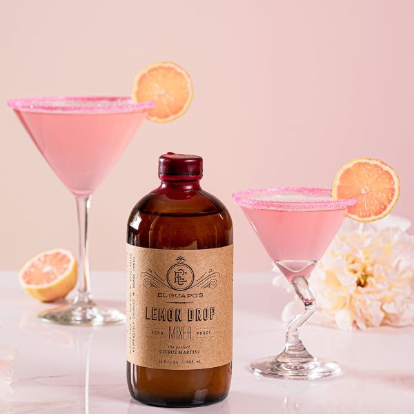 A bottle of El Guapo Lemon Drop Mixer next to two pink Cosmopolitan cocktails.