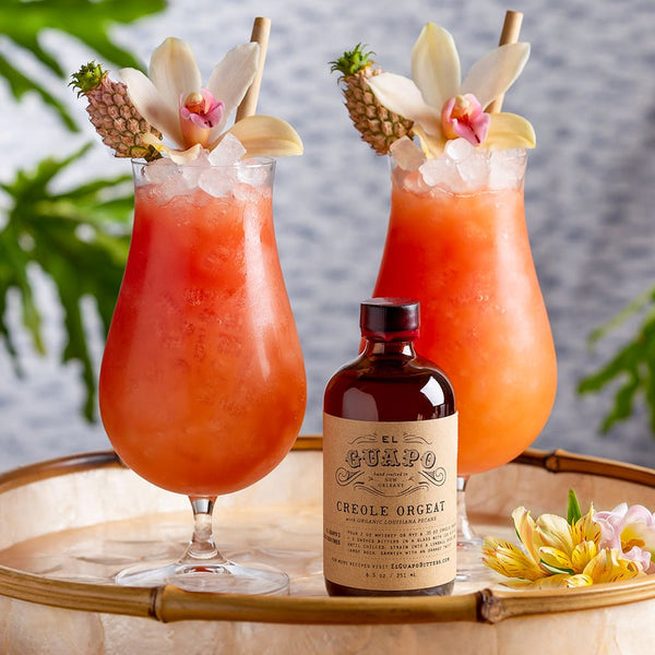 Two tropical cocktails with decorative elements and a bottle of small Gumpo Creole Orgeat on a wooden tray.