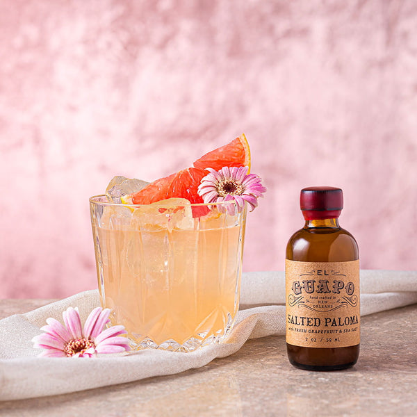 Paloma Cocktail with grapefruit and flowers next to a bottle of El Guapo Single Serving Salted Paloma Mixer