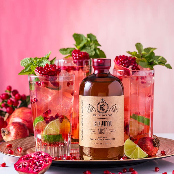 El Guapo Mojito Mixer styled with pomegranate, mint, and lime garnishes on a pink background.