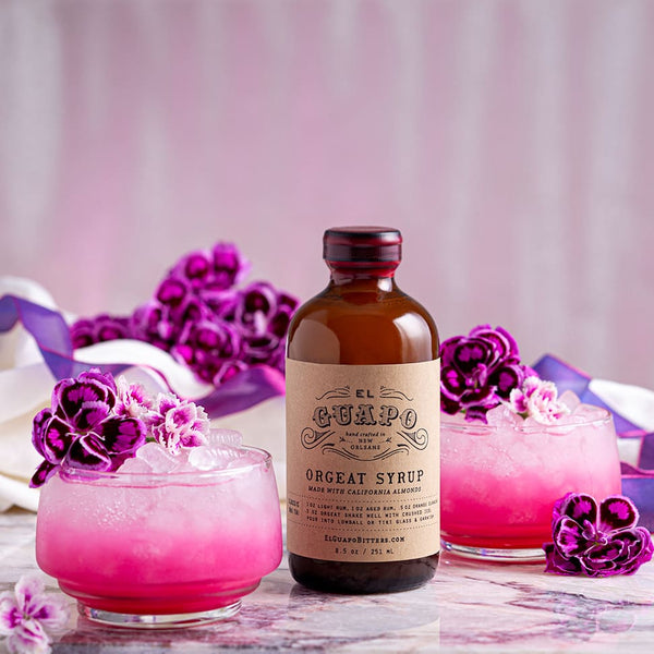 Bottle of small 8.5 oz El Guapo Orgeat Syrup with pink cocktails and flowers on a marble surface.