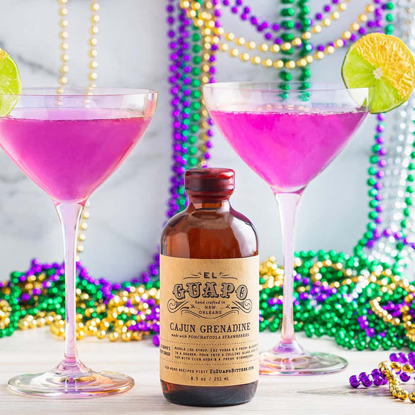 Two purple cocktails with gold dusted lime wheel garnishes in purple tinted martini glasses with a bottle of El Guapo Cajun Grenadine, surrounded by Mardi Gras beads.