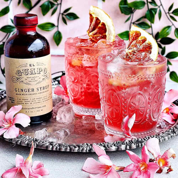 Two pink cocktails with garnishes on a silver tray next to a bottle of El Guapo Ginger Syrup.