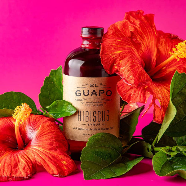Small 8.5 oz bottle of El Guapo Hibiscus Syrup surrounded by red hibiscus flowers on a pink background