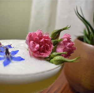 Close-up of a foamy cocktail garnished with pink roses and small blue edible flowers