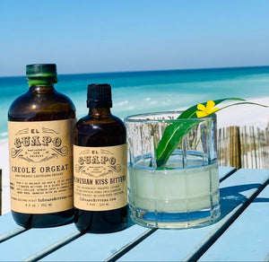 El Guapo Creole Orgeat and Polynesian Kiss Bitters bottles next to a beachside cocktail