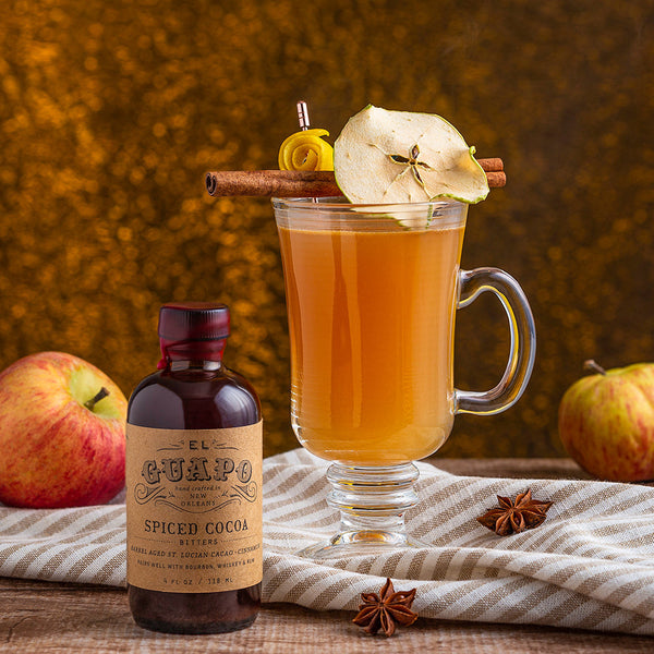 Spiked Hot Cider with a cinnamon stick garnish next to El Guapo Spiced Cocoa Bitters