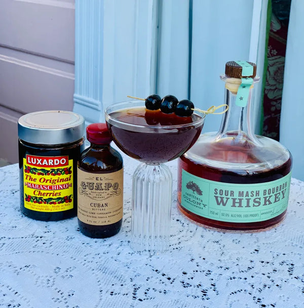 The Graveyard Shift cocktail next to El Guapo Cuban Bitters and Bourbon Cherry Syrup