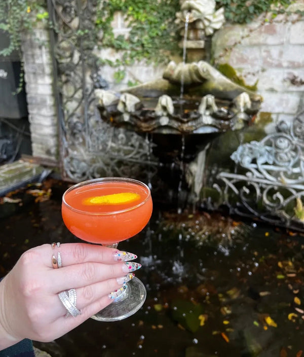 The Kensington cranberry orange cocktail in a coupe glass held by a hand in front of a fountain