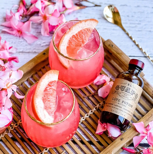 Two pink grapefruit cocktails on a tray with a bottle of El Guapo Love Potion Bitters