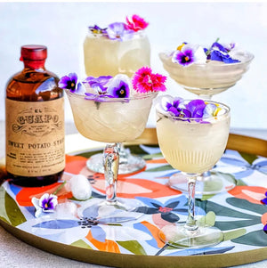 Mardi Gras Margaritas with purple edible flowers next to a bottle of El Guapo Syrup