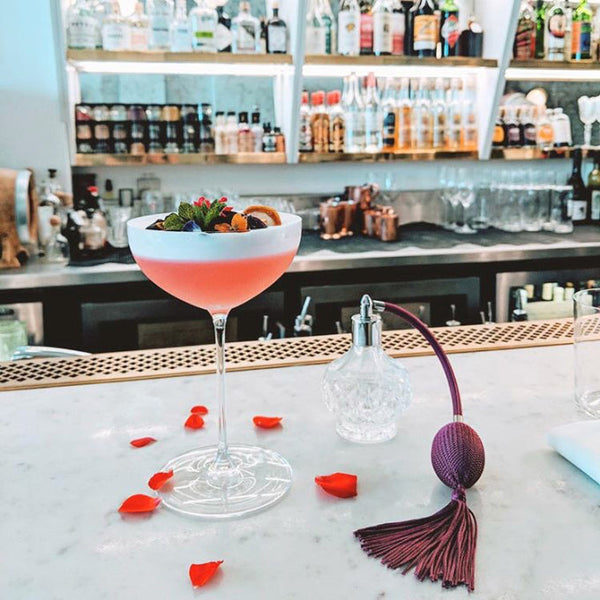 Pink Elephant gin cocktail with rose petals and a vintage glass perfume atomizer on a bar
