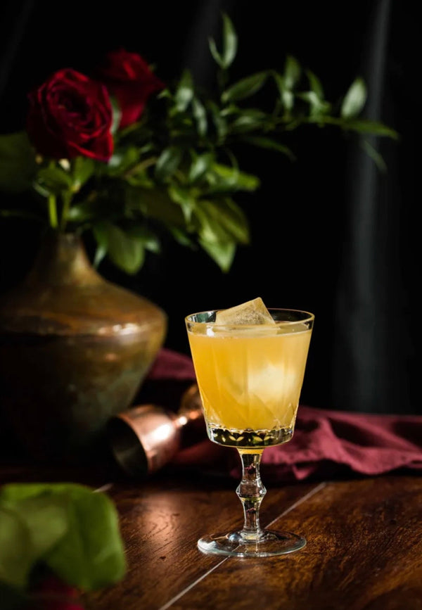 Rose Ward 23 cocktail in a stemmed glass on a wooden table next to a vase of red roses