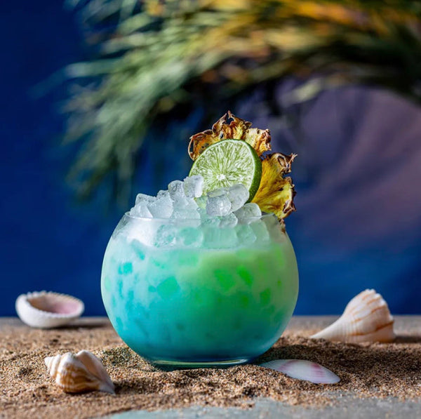 Blue Tidal Wave gin tiki cocktail with crushed ice, lime, and dried pineapple on sand