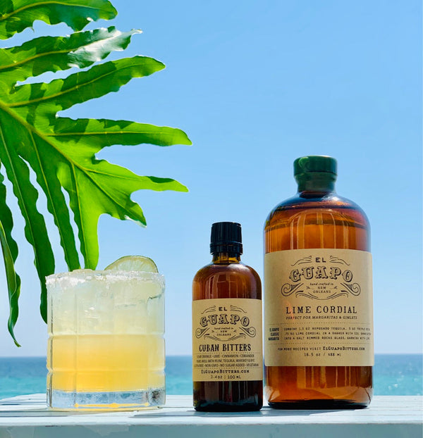 Margarita cocktail next to El Guapo Cuban Bitters and Lime Cordial on a beach table