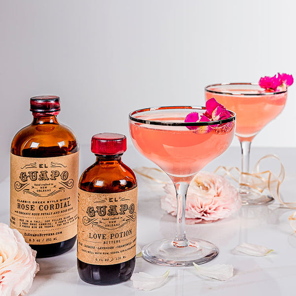 An 8.5 oz bottle of El Guapo Rose Cordial and bottle of El Guapo Love Potion Bitters next to two pink cocktails garnished with pink flowers on a light background.