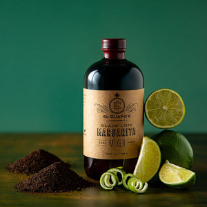 Bottle of El Guapo Black Lime Margarita Mixer styled with fresh limes and black lime powder on a wooden surface with a green background
