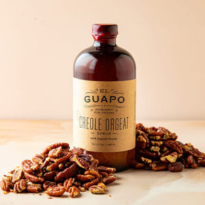 Bottle of  large Guapo Creole Orgeat syrup  surrounded by pecans on a wooden surface