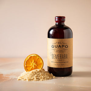 Bottle of large 16.5 oz El Guapo Demerara syrup with a slice of orange and sugar on a beige background