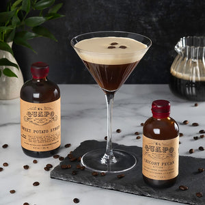 Espresso Martini with espresso bean garnish, El Guapo sweet potato syrup bottle, and chicory pecan bitters bottle on a marble surface decorated with espresso beans