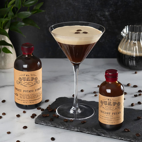Espresso Martini with espresso bean garnish, El Guapo sweet potato syrup bottle, and chicory pecan bitters bottle on a marble surface decorated with espresso beans