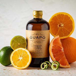 Large bottle of El Guapo Tonic Syrup with citrus fruits on a light background
