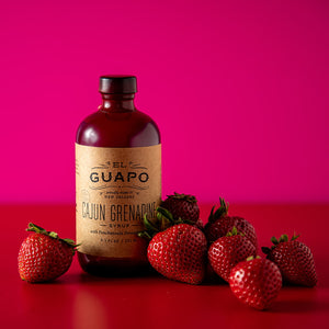 8.5 oz Bottle of Guapo Cajun Grenadine syrup with strawberries on a red surface and pink background