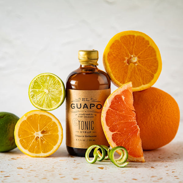 Bottle of small El Guapo Tonic with sliced oranges and limes on a light background