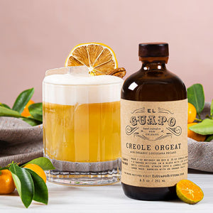 Bottle of small El Guapo Creole Orgeat with a cocktail garnished with a dehydrated lemon wheel on a light background with kumquats