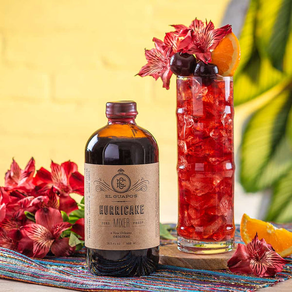 Bottle of El Guapo Hurricane Mixer with a Hurricane cocktail garnished with flowers, cherries and oranges on a colorful tablecloth.