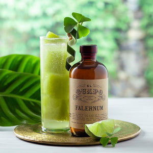Bottle of El Guapo Falernum with a glass filled with a green cocktail on a gold plate with a blurred green background