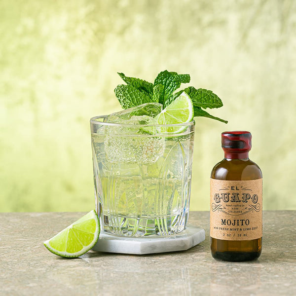 Mojito cocktail with lime wedges and mint leaves next to a bottle of Single Serving El Guapo Mojito Mixer on a coaster atop a wooden surface.