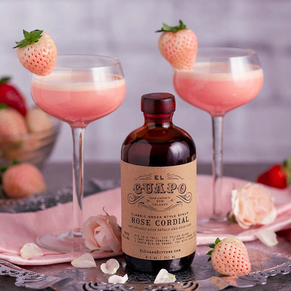 Bottle of El Guapo Rose Cordial with two creamy pink cocktails garnished with light pink strawberries on a decorative table.