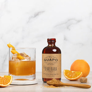 Bottle of small 8.5 oz Guapo Demerara syrup with a cocktail, sugar and oranges on a marble surface
