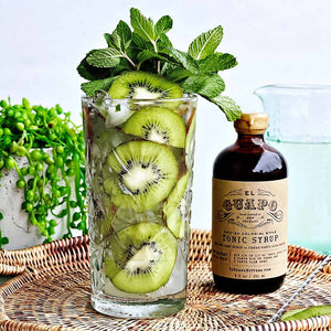 Glass with cocktail and kiwi slices as garnishes with mint leaves and a bottle of El Guapo tonic syrup on a woven tray.