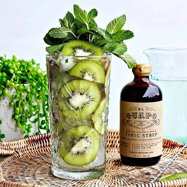 Glass with cocktail and kiwi slices as garnishes with mint leaves and a bottle of El Guapo tonic syrup on a woven tray.