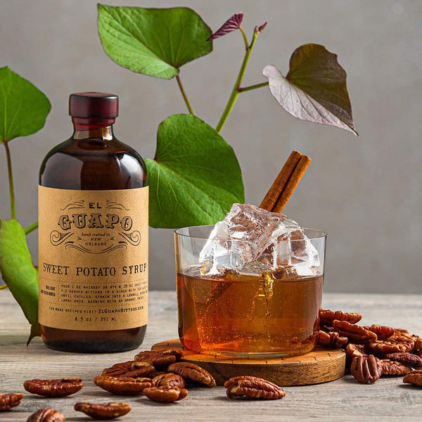 Small bottle of El Guapo sweet potato syrup with an old fashioned garnished with a cinnamon stick surrounded by whole pecans on a wooden surface.