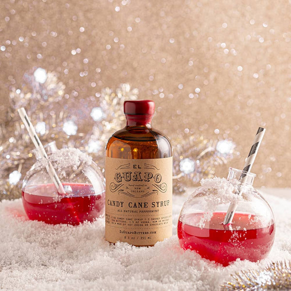 Bottle of El Guapo Candy Cane Syrup with Christmas cocktails poured into plastic ornaments on a snowy background