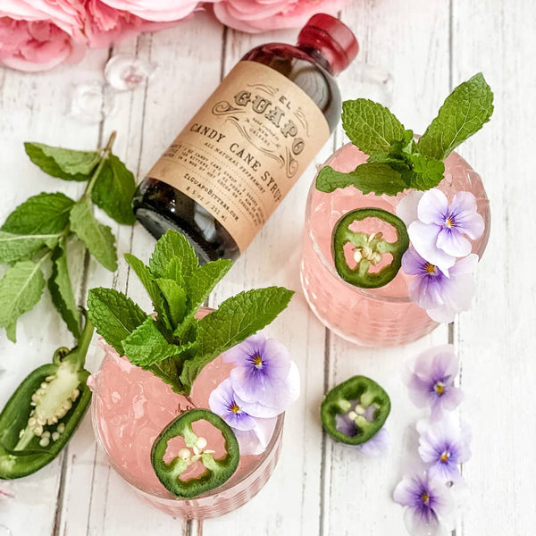 Two pink cocktails with jalapeño slices and flowers, a bottle of El Guapo candy cane syrup on a wooden surface.
