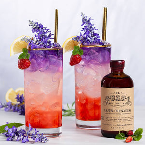 Two layered colorful cocktails with miniature strawberry, lemon wedge and flower garnishes and a bottle of El Guapo Cajun Grenadine on a light background