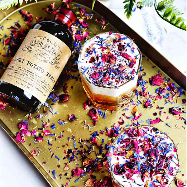 Two glasses filled with a colorful drink and sweet potato syrup bottle on a decorative tray garnished with dried flower petals