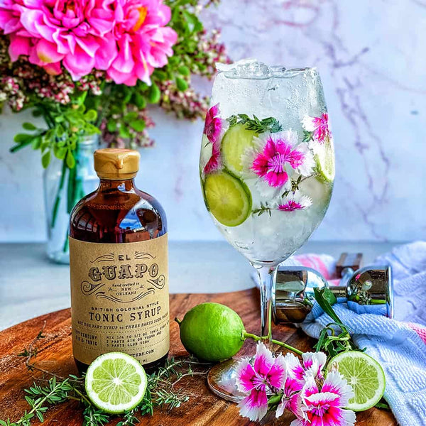 Gin and tonic with floral garnish, bottle of El Guapo Tonic Syrup, and fresh limes on a wooden board.